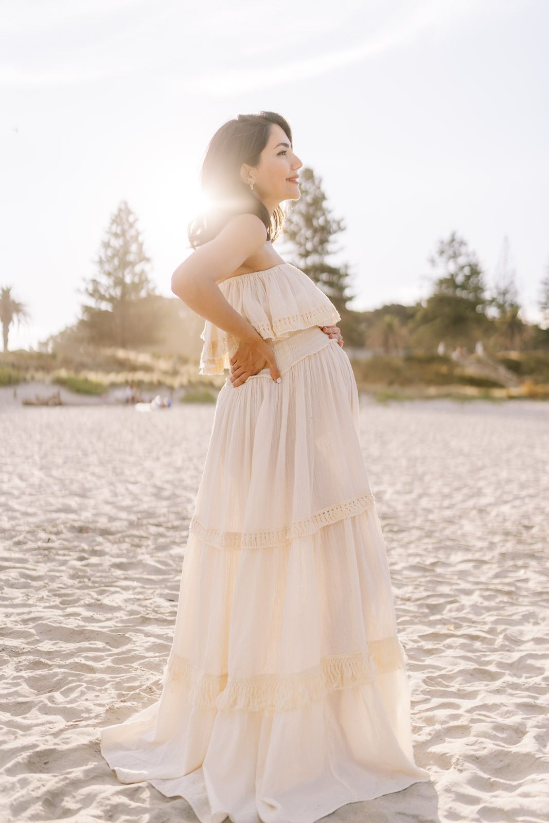 Woman in cream boho maxi dress standing on sandy beach with golden backlight sunset glow behind her.
