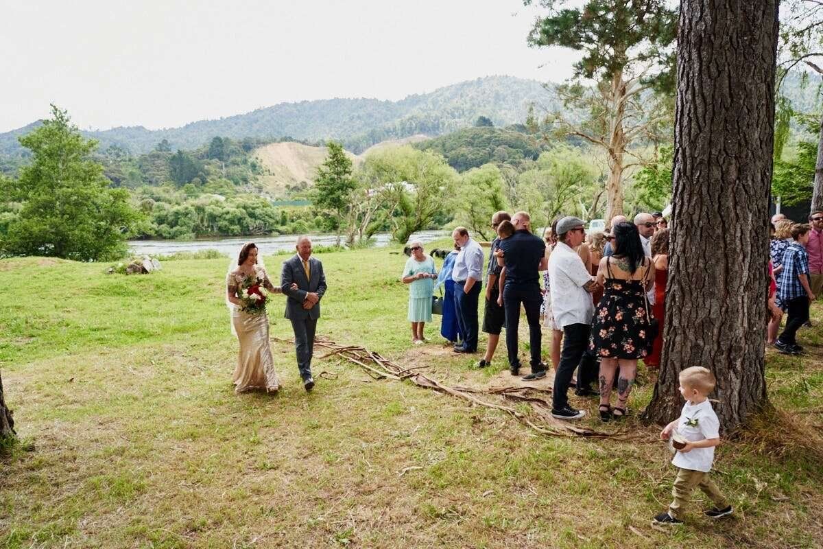 Outdoor wedding ceremony on green meadow with guests gathering near trees, river and forested hills in background.