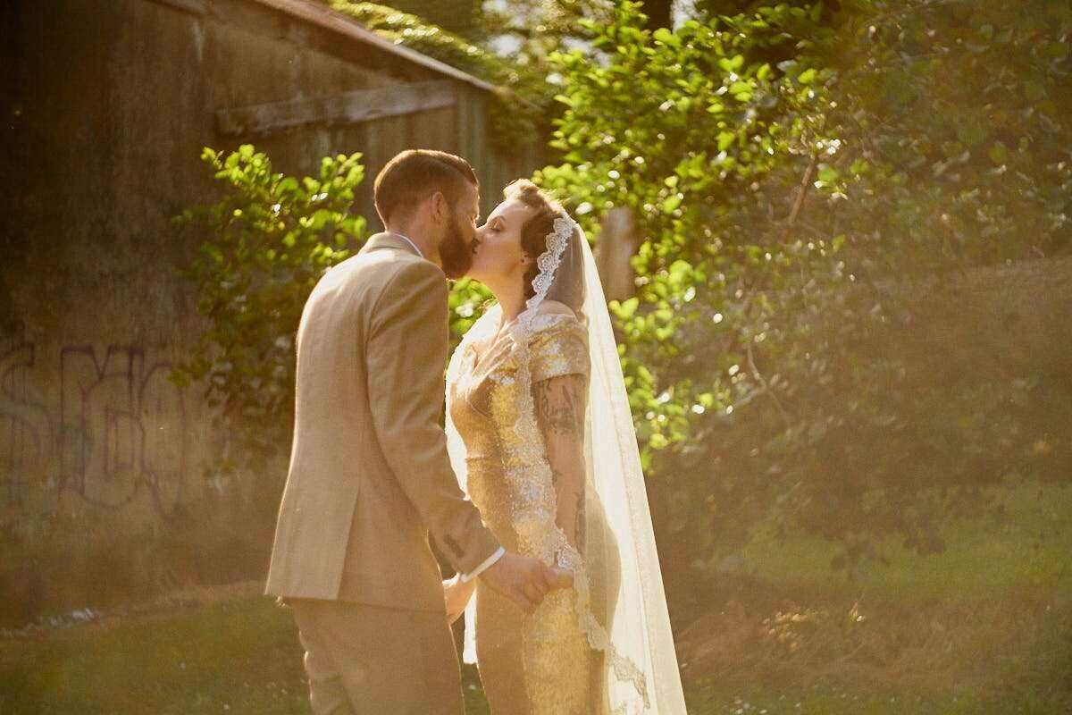 Bride and groom sharing a romantic kiss outdoors in golden sunlight, bride wearing a lace gown and veil.