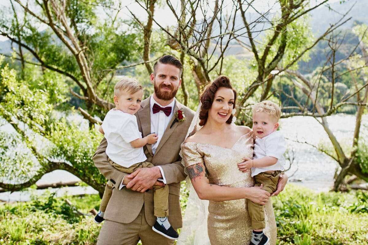 A smiling family in formal attire poses outdoors, with a bearded groom, elegant bride, and two young boys amid lush greenery.