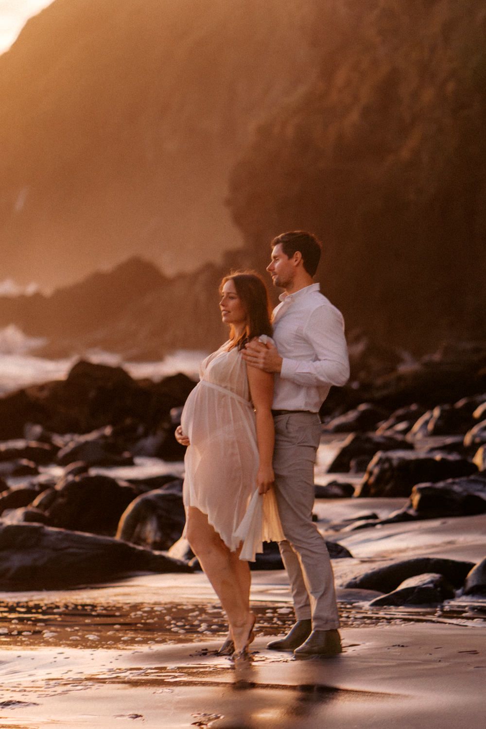 Couple embracing on a rocky beach at golden sunset, pregnant woman in white dress with partner in white shirt.