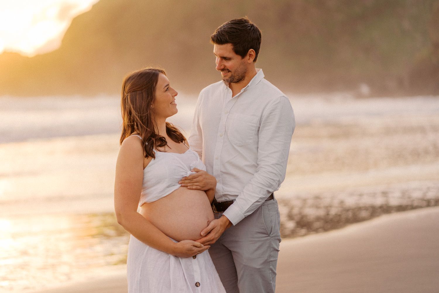 Pregnant couple embracing on beach at golden sunset, woman in white dress cradling baby bump.