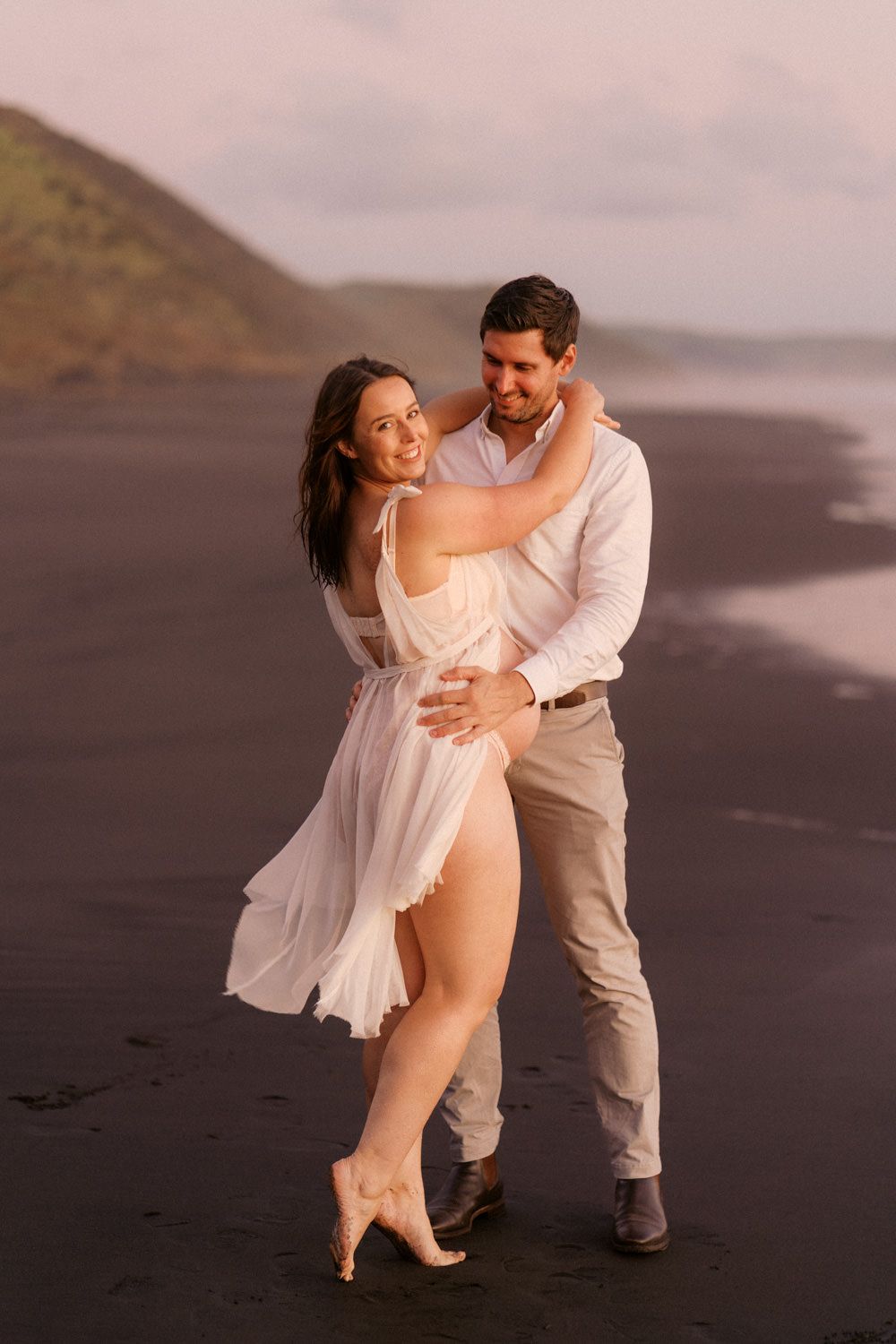 Couple embracing on a black sand beach at sunset, woman in white flowing dress, man in white shirt and khaki pants.
