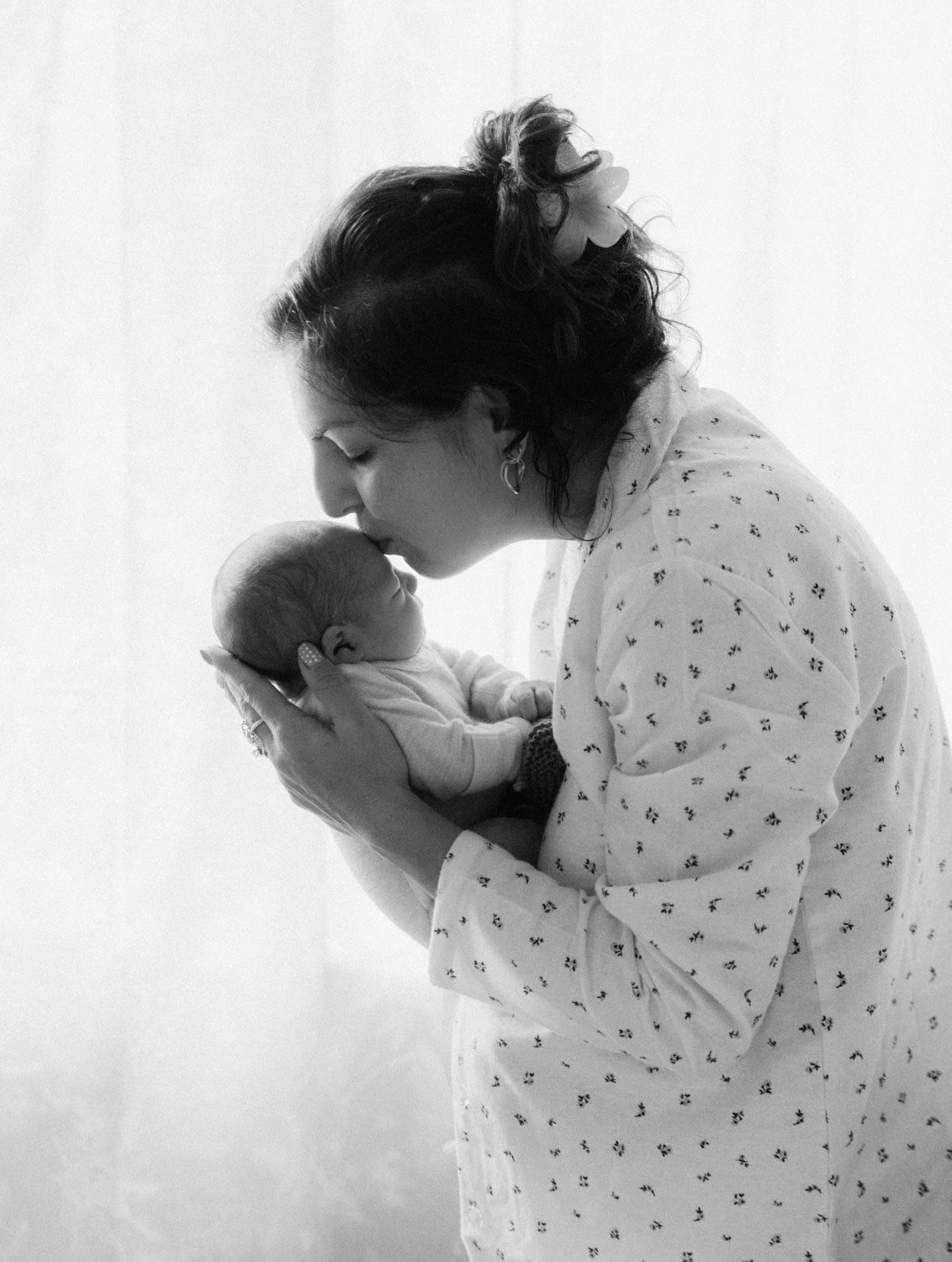 Black and white photo of a mother in floral robe kissing her newborn baby held gently in her hands.