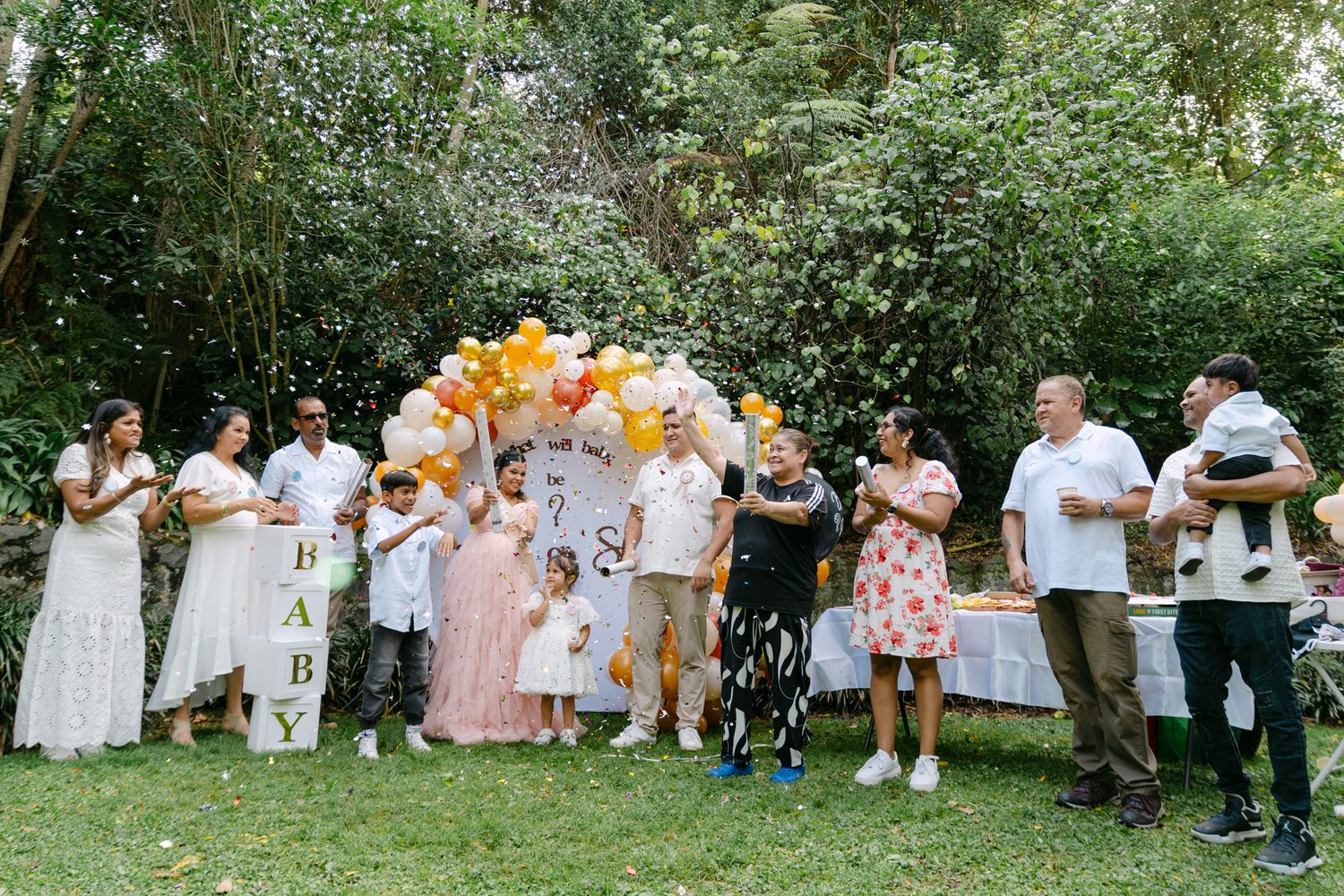 Outdoor baby shower party with guests, yellow and white balloons, and a BABY sign in a lush garden setting.