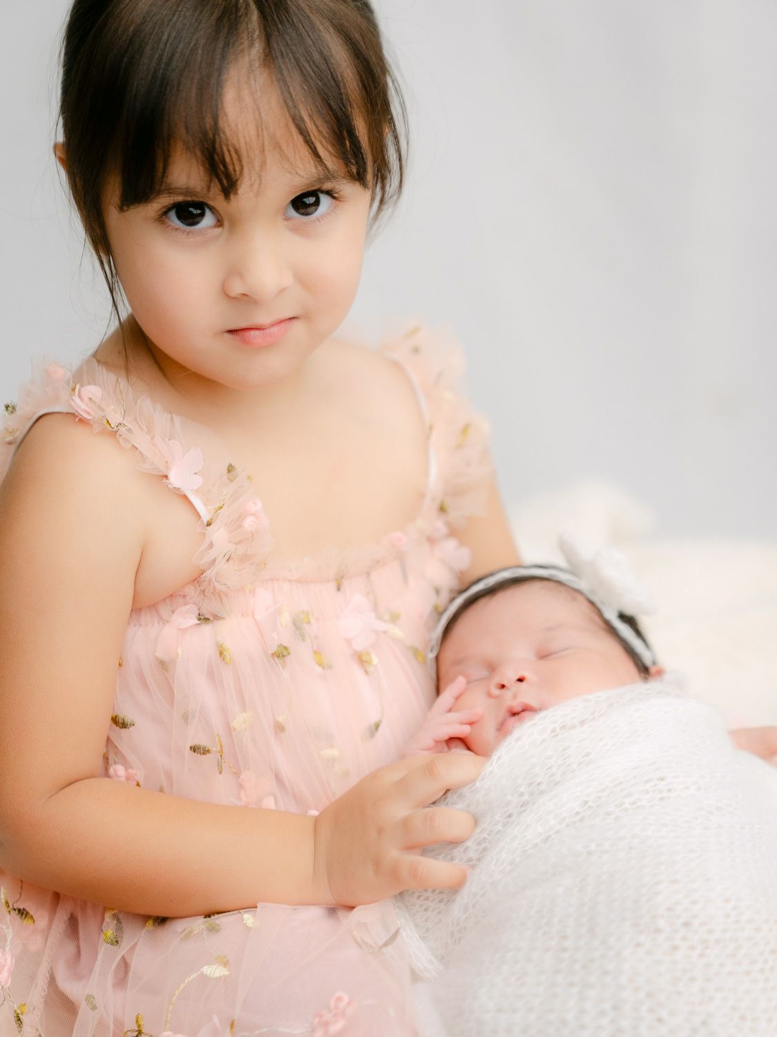 A young girl in a pink sequin dress tenderly holds a sleeping newborn baby wrapped in white.