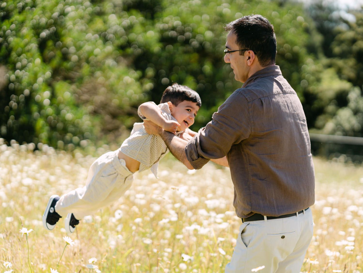 Father joyfully swinging his young son in a sunny wildflower meadow, sharing a playful outdoor moment.