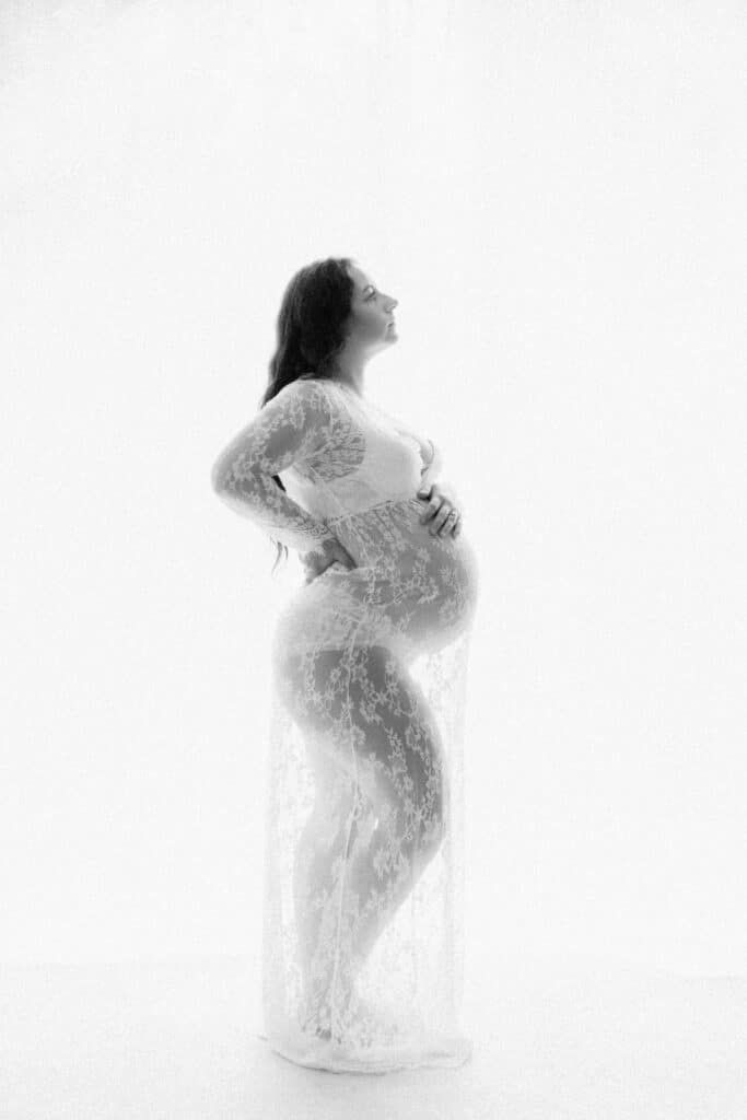 Black and white maternity photo of pregnant woman in lace gown cradling baby bump against white background.