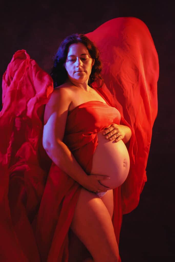 Pregnant woman draped in flowing red fabric poses gracefully against dark background, cradling her baby bump.