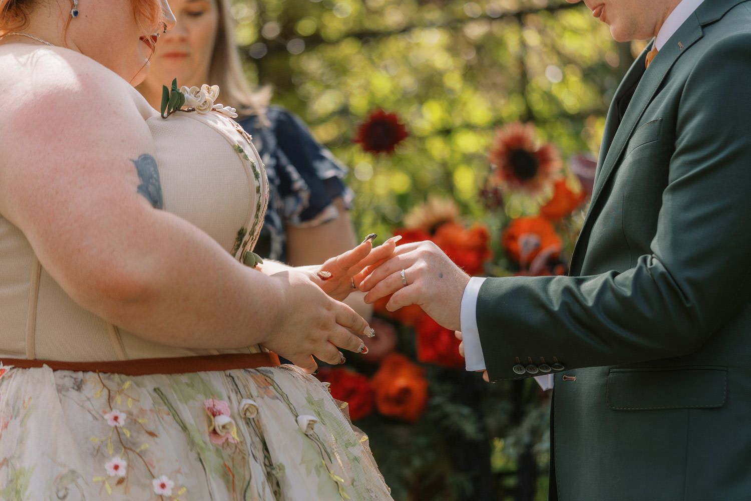Couple exchanging rings during outdoor wedding ceremony with colorful floral backdrop.