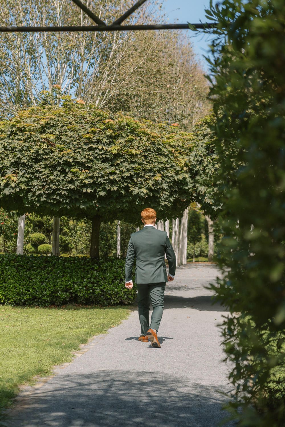 A red-haired man in a green suit walks along a garden path lined with trimmed hedges and trees on a sunny day.