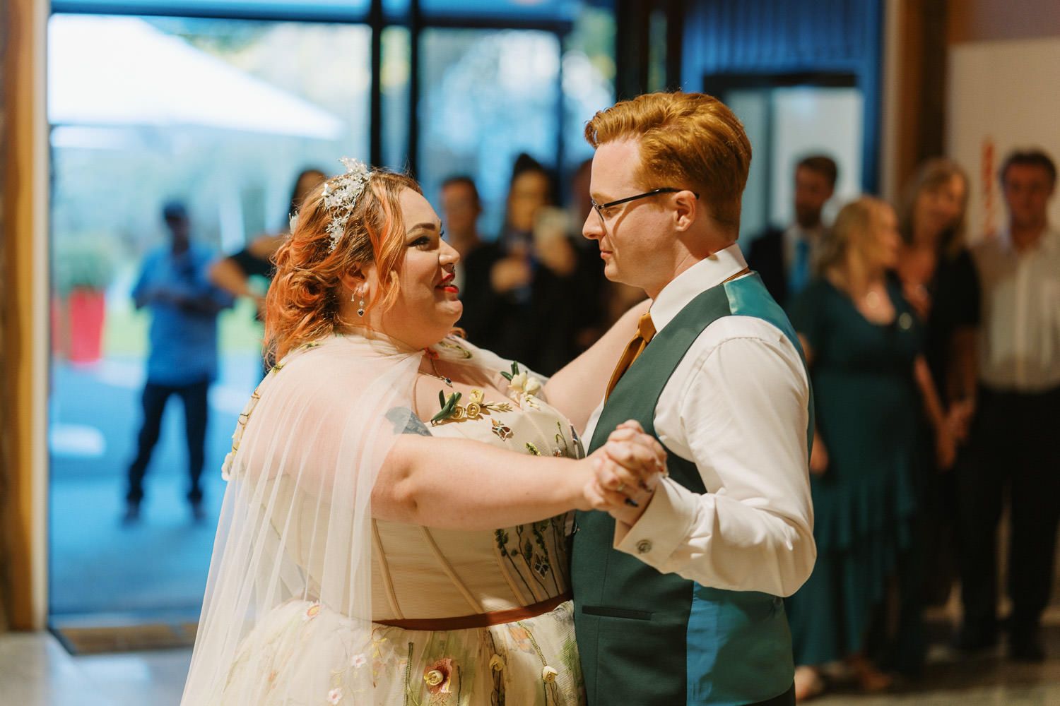 Bride and groom share their first dance at wedding reception with guests watching in background.