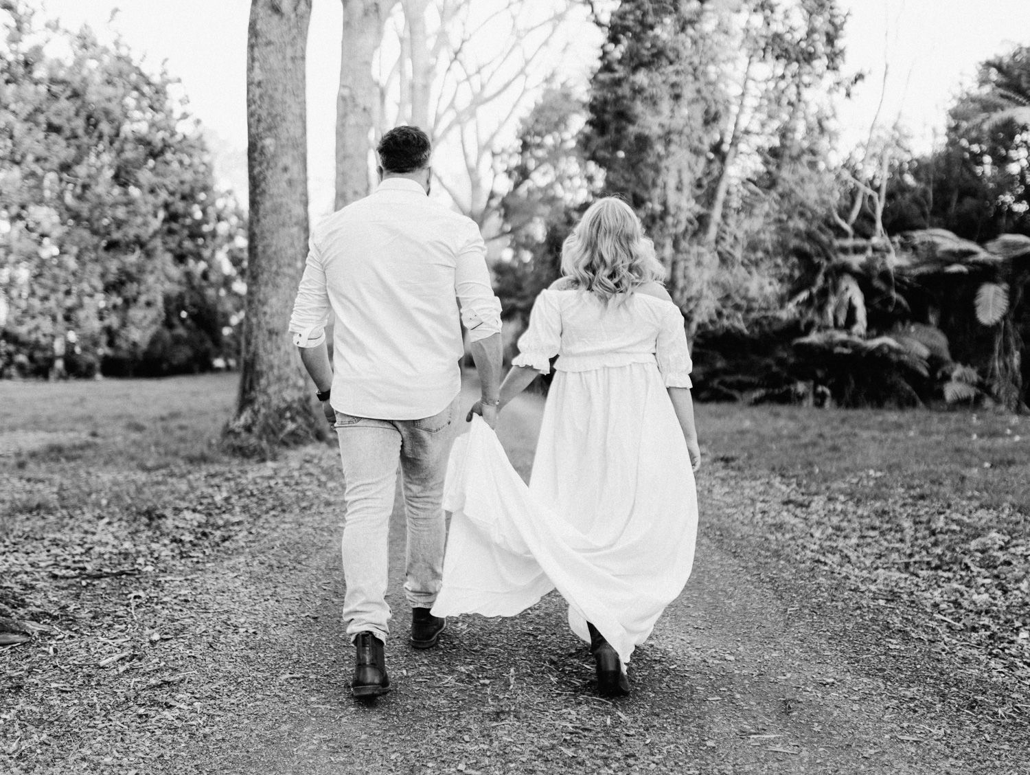 Black and white photo of a couple holding hands walking down a garden path, woman in flowing white dress.