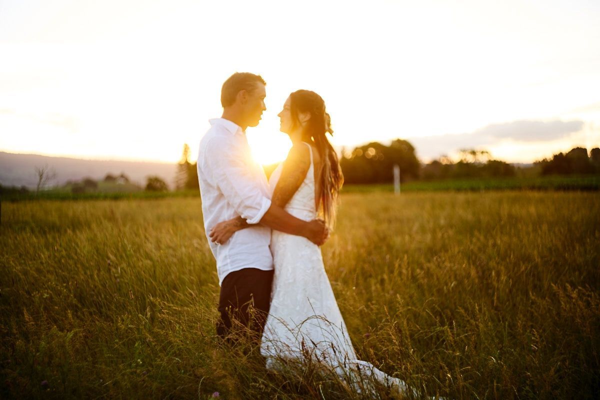 Couple embracing in golden sunset light in a lush green field, wearing white wedding attire.