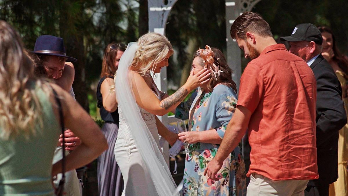 Bride in white gown and veil shares emotional moment with guest at outdoor wedding ceremony on sunny day.