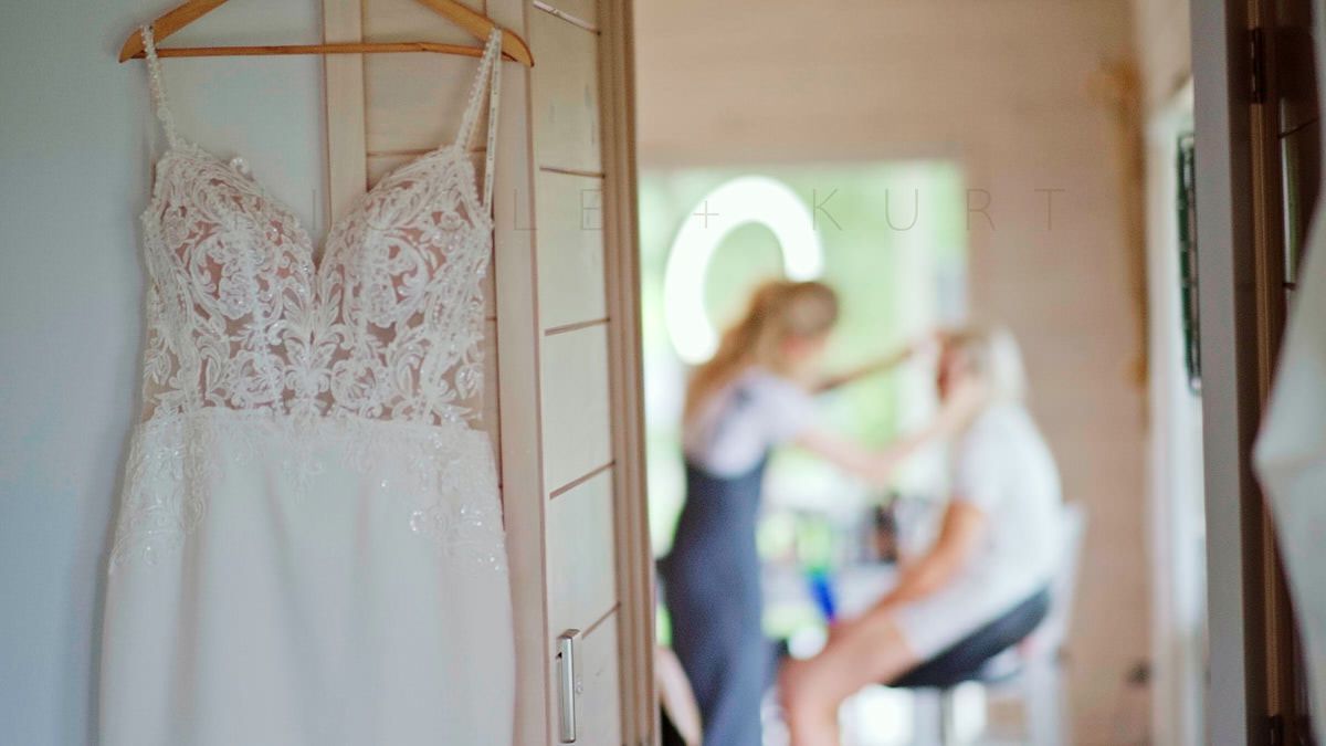 Wedding dress on hanger in foreground with bridal party getting ready in soft-focus background.