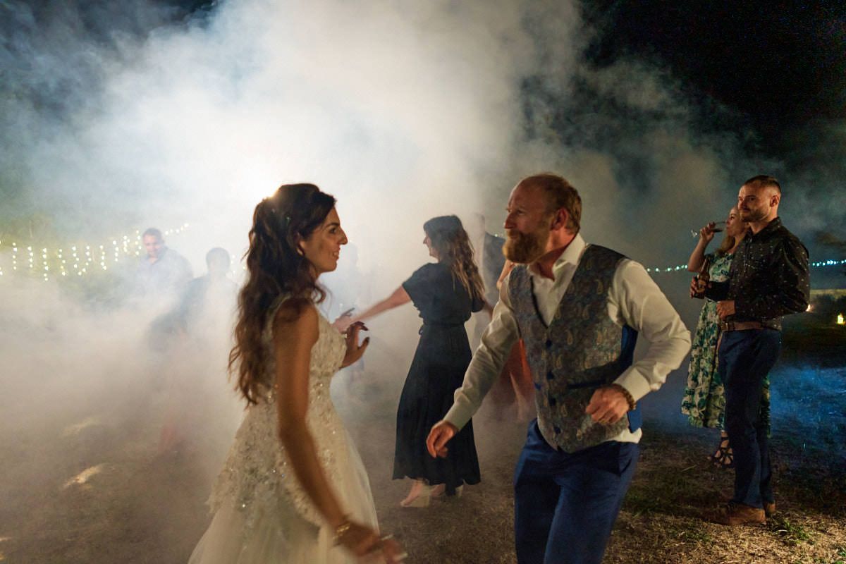 Bride and groom dancing joyfully in dramatic fog at an outdoor evening wedding reception.