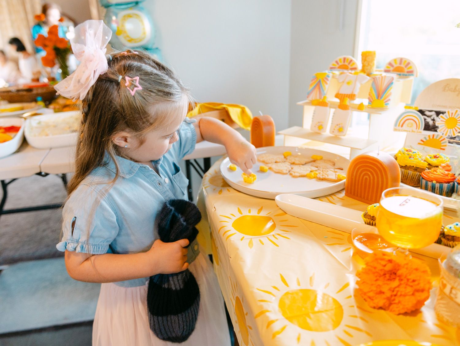 Young girl in blue dress decorating cookies at a sunshine-themed yellow party table with cupcakes and baking supplies.