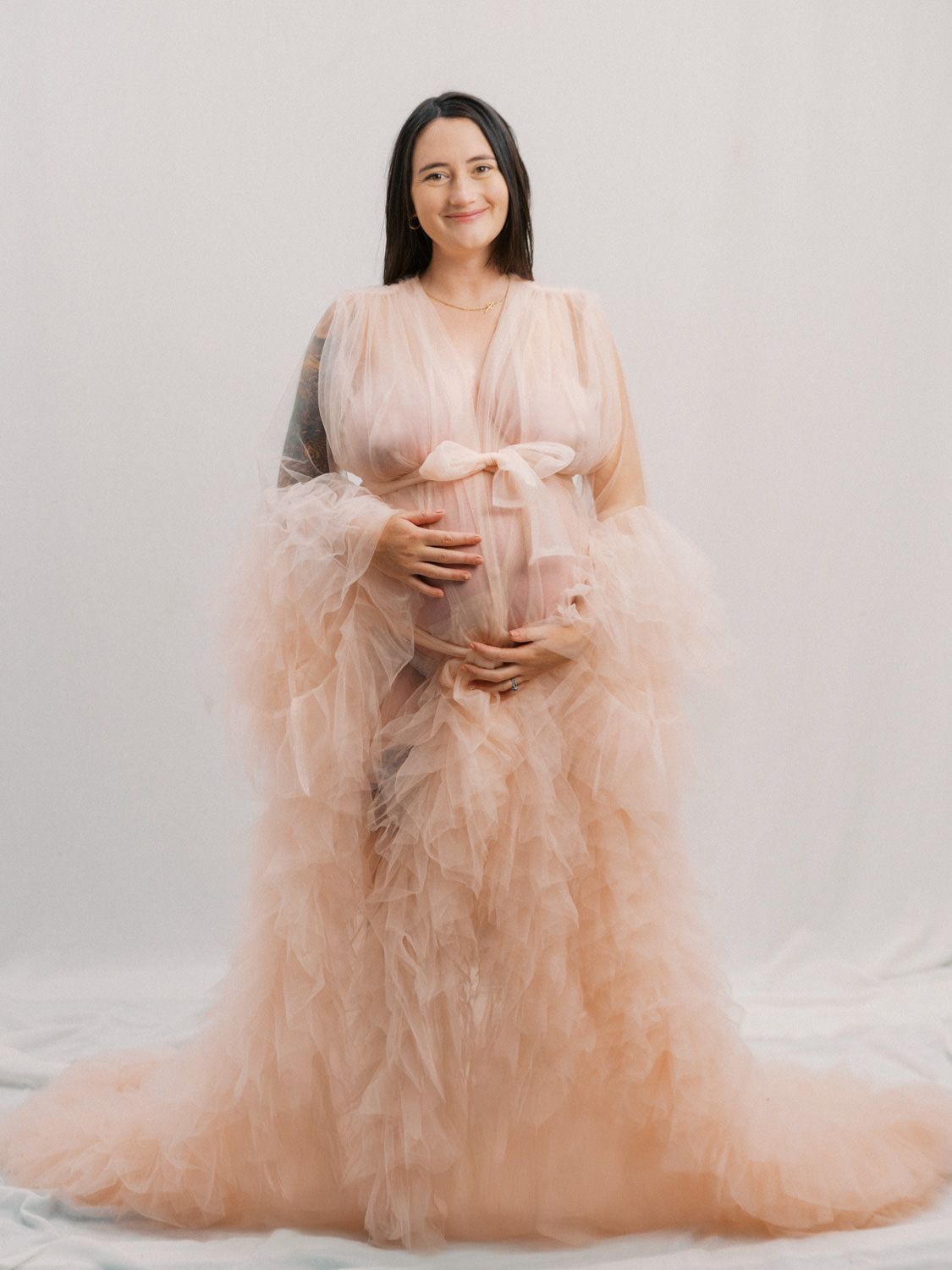 Pregnant woman in elegant peach tulle maternity gown cradling baby bump during professional studio photo session.