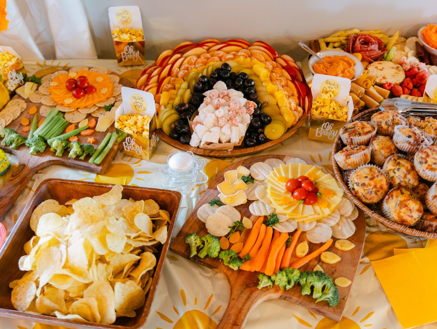 Colorful party spread with chips, veggie platters, fruit arrangements, and appetizers on a sunny yellow tablecloth.