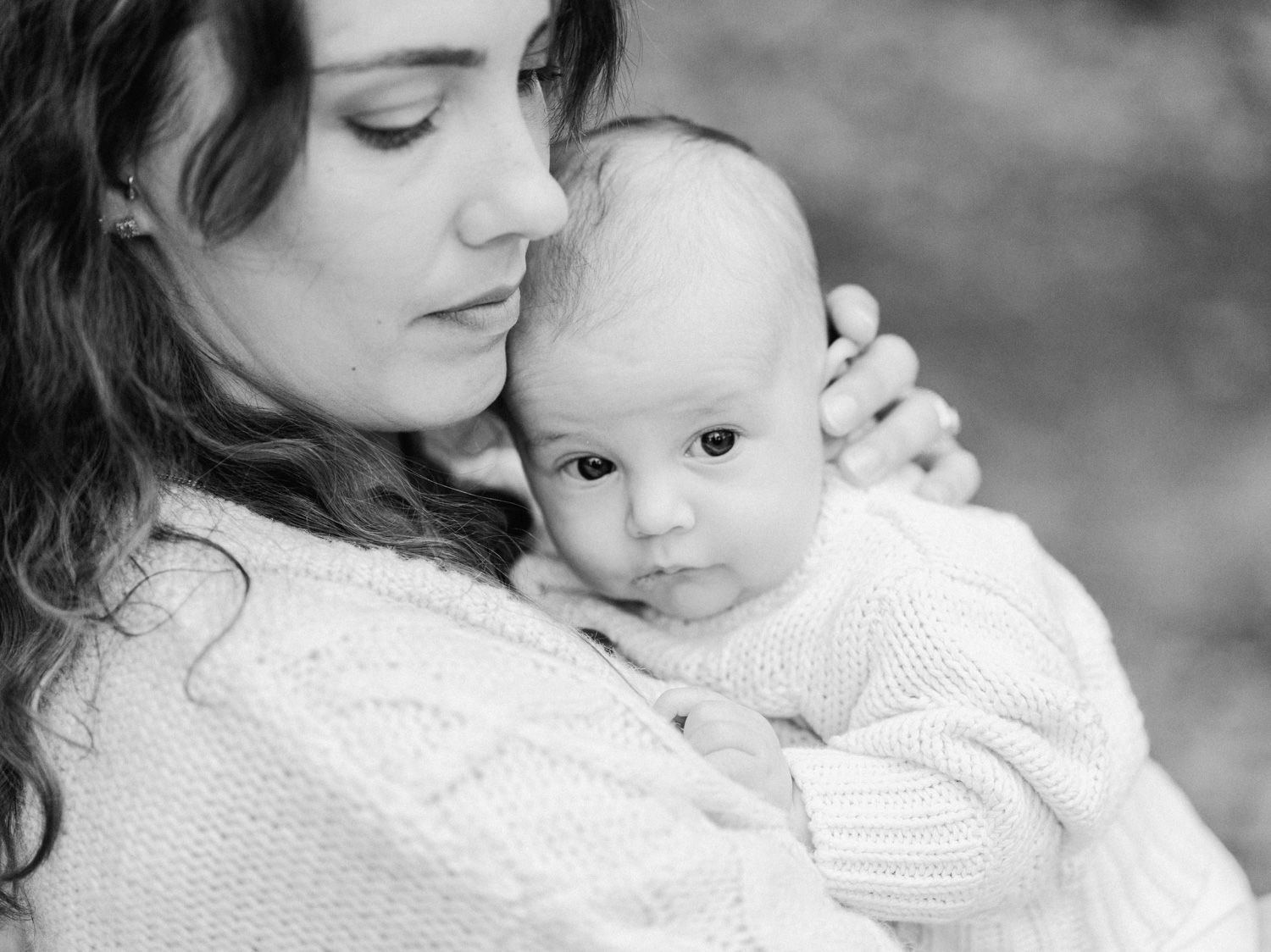 Black and white portrait of a mother tenderly holding her newborn baby in a cozy knit sweater outdoors.