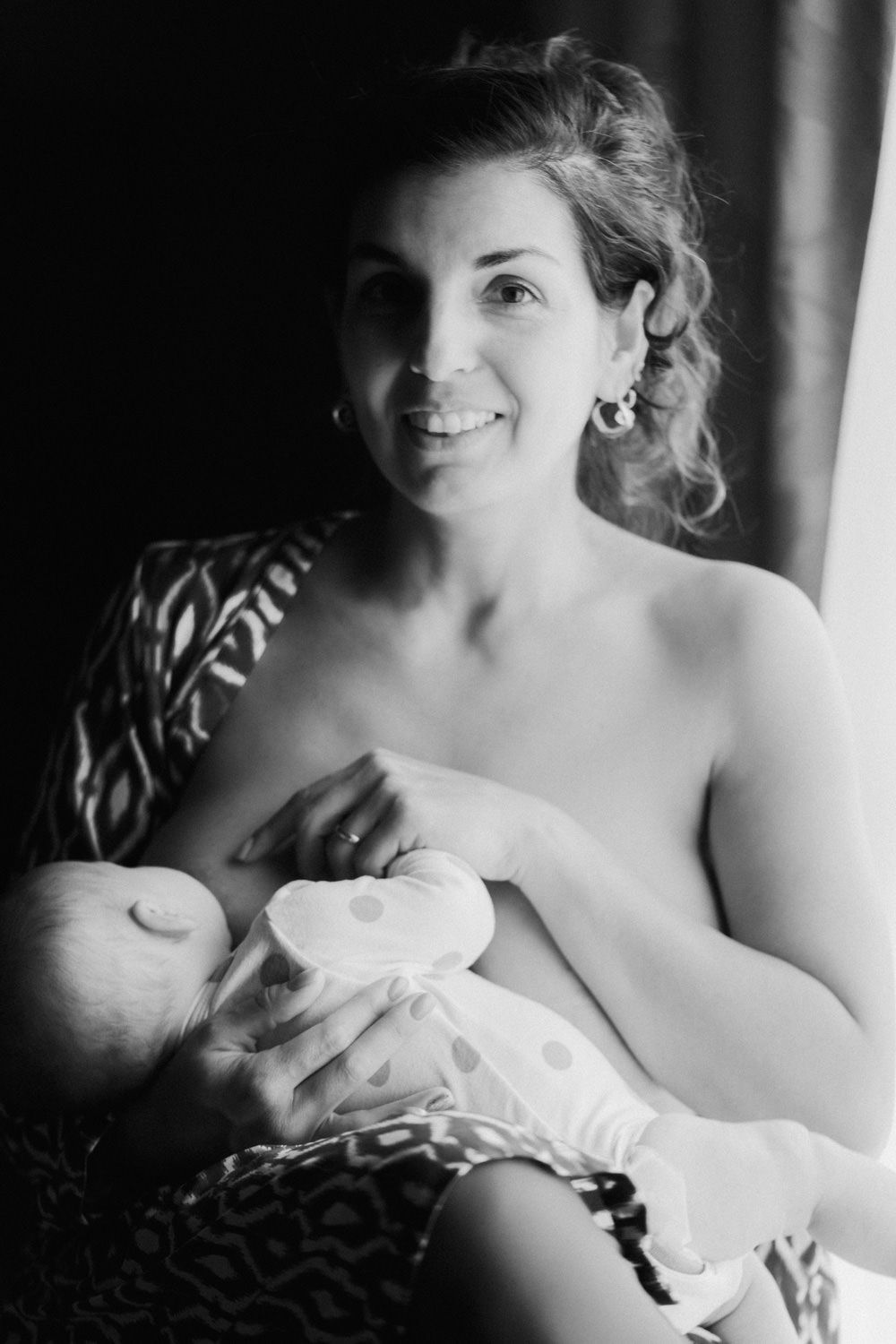 Black and white photo of a smiling mother breastfeeding her newborn baby while seated in a patterned chair.