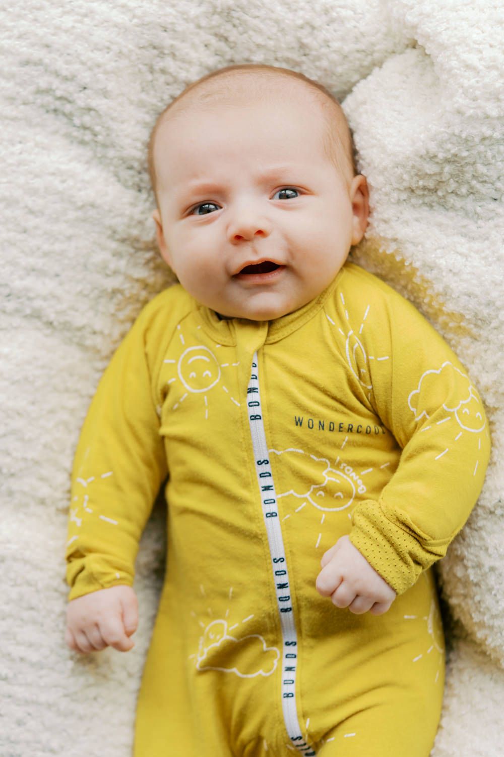 A smiling newborn baby wearing a yellow Bonds Wondercool zipsuit with sun and cloud prints, lying on a white fluffy blanket.