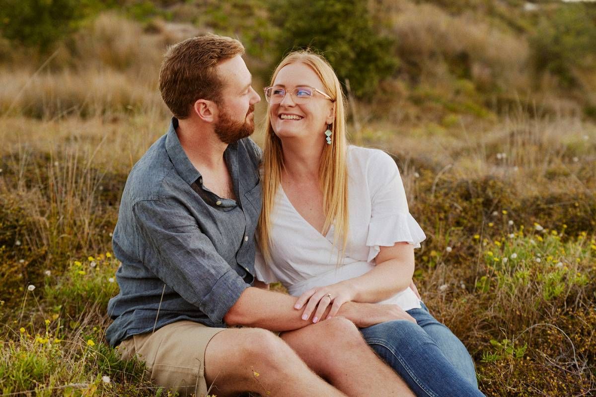 Couple sitting in a sunny meadow, woman laughing as man leans in close, wearing casual summer outfits.