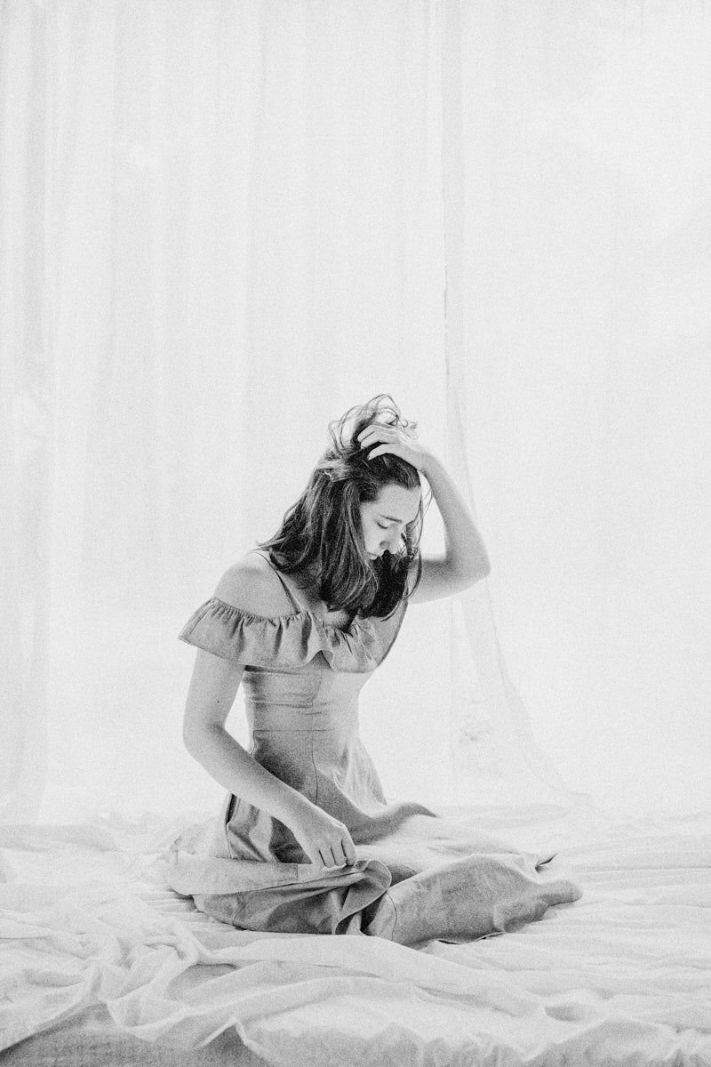Black and white photo of a woman in an off-shoulder dress sitting cross-legged on a bed, hand in hair.