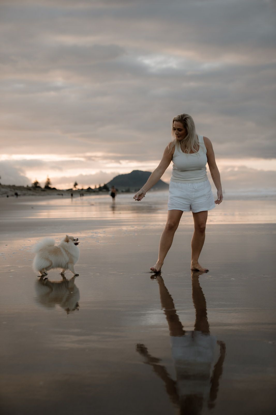 Woman in white outfit playing with a fluffy white Spitz dog on a reflective beach at sunset.
