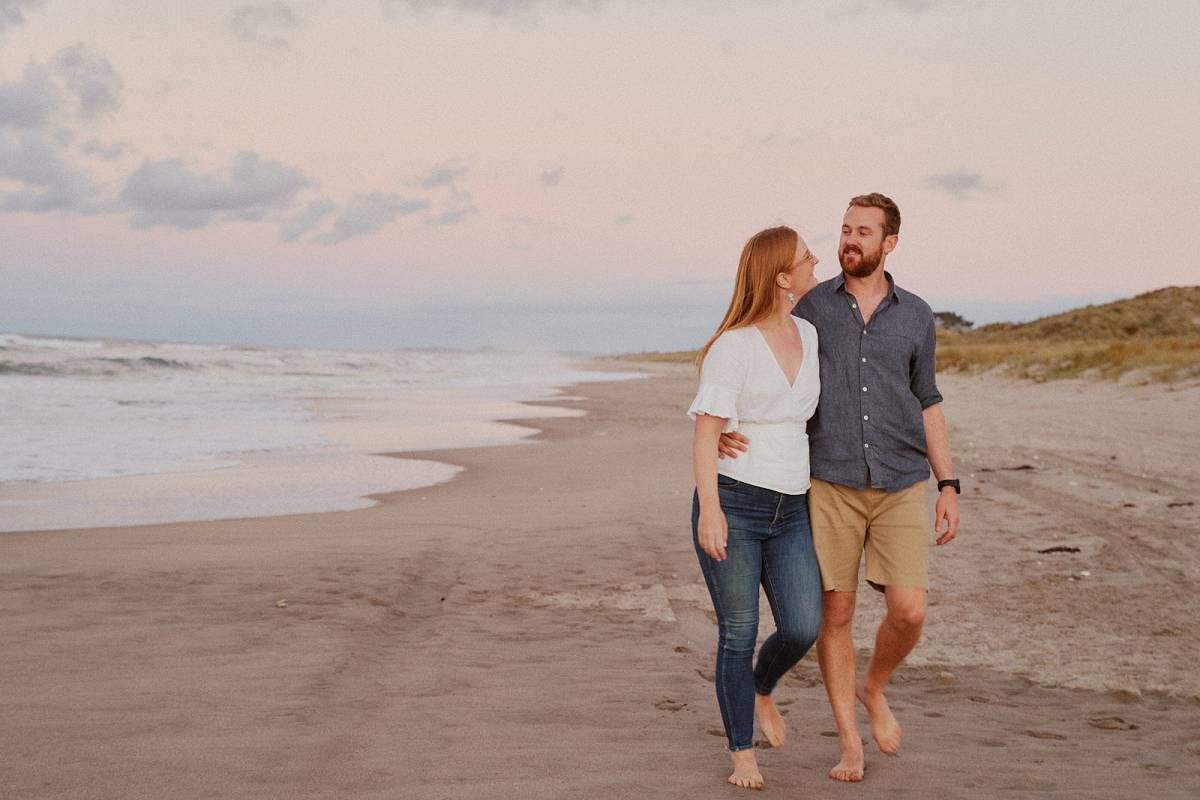 Couple walking barefoot on a beach at sunset, smiling at each other near the ocean.