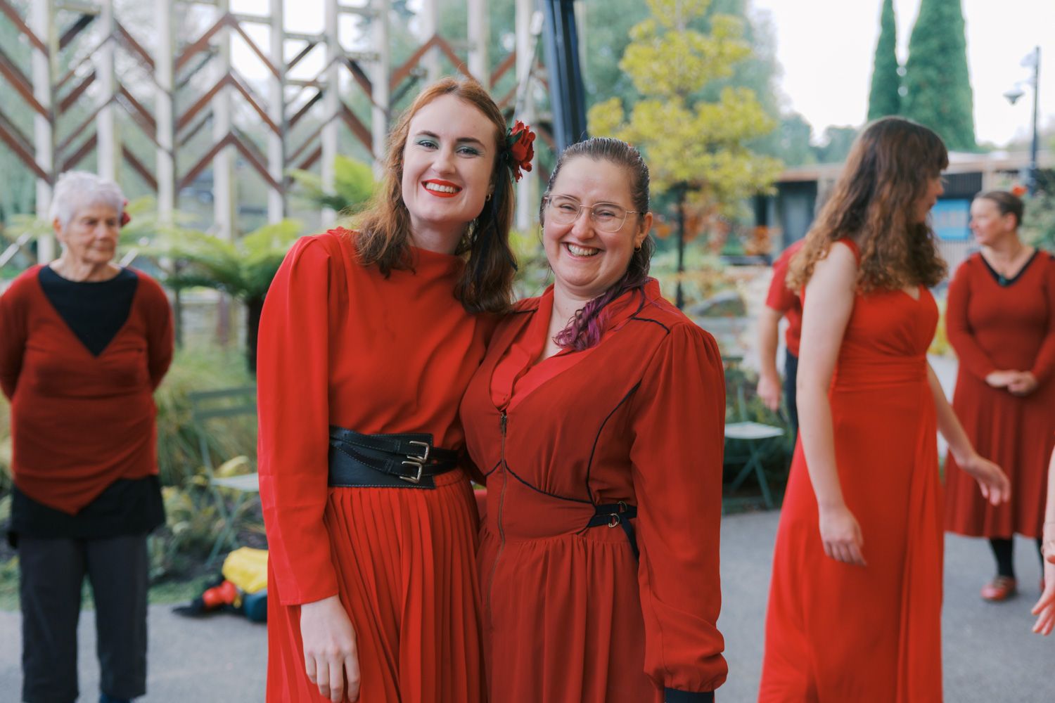 Two women in red dresses smile together, with a choir group in matching red outfits visible in the background.