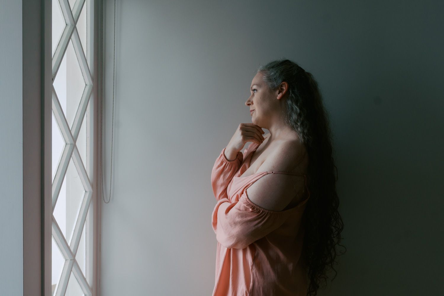 Woman with curly hair gazing thoughtfully out a window in soft pink dress, bathed in natural light.