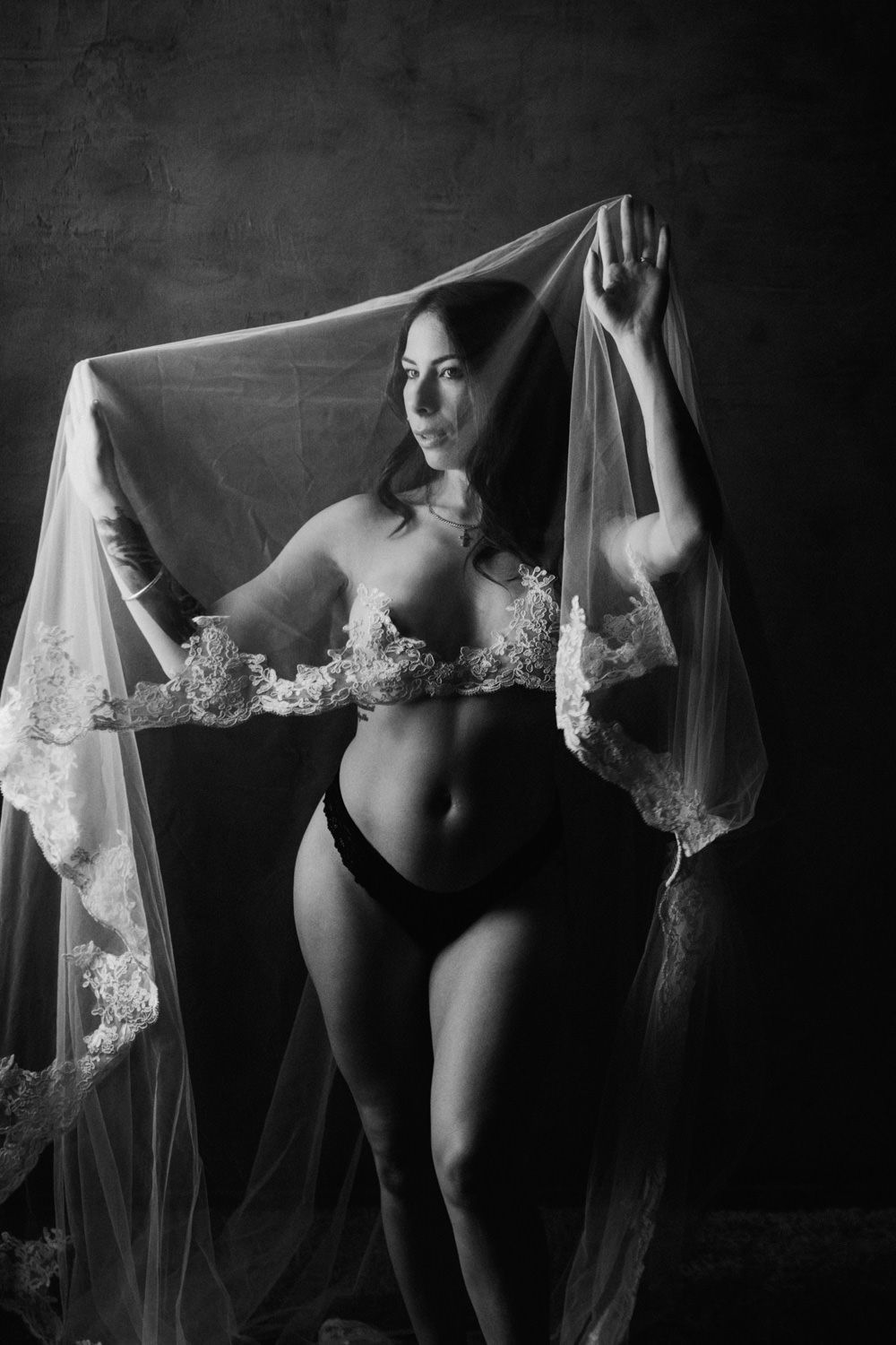 Black and white boudoir photo of a woman in lace lingerie holding a sheer veil overhead in a studio setting.