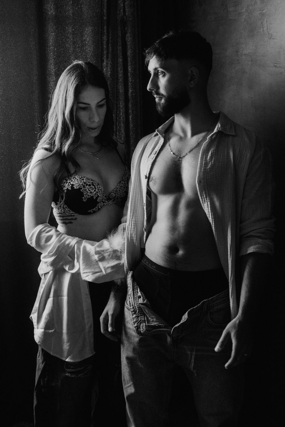 Black and white photo of a couple posing intimately, man with open shirt showing abs, woman in lace bra and white shirt.