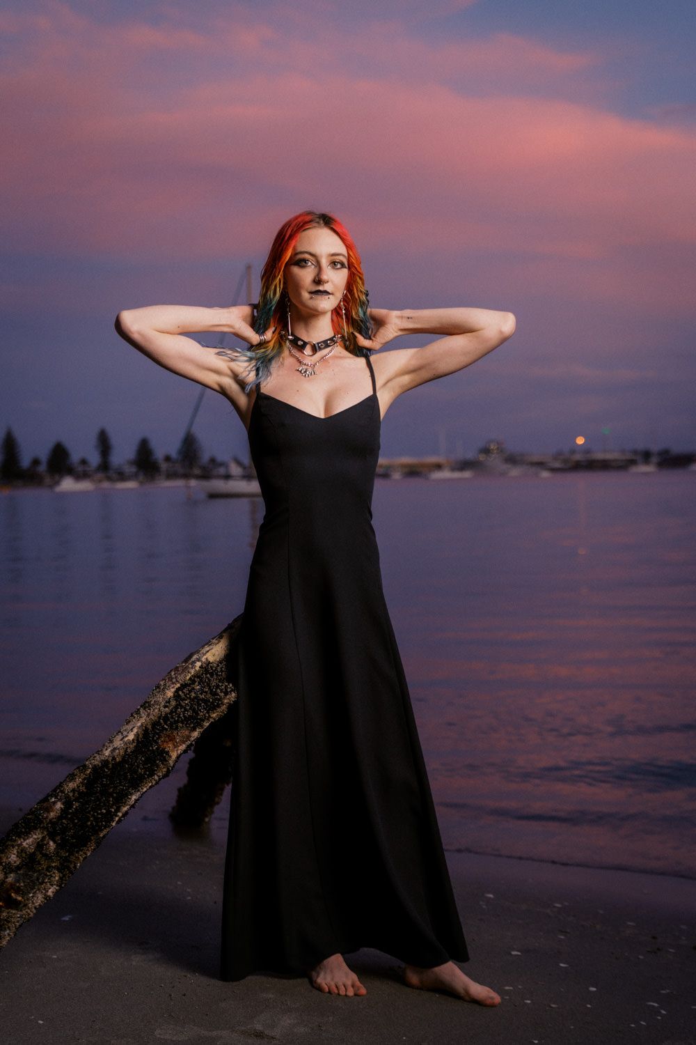 Woman in black maxi dress poses barefoot by waterfront at sunset with colorful pink and purple sky backdrop.