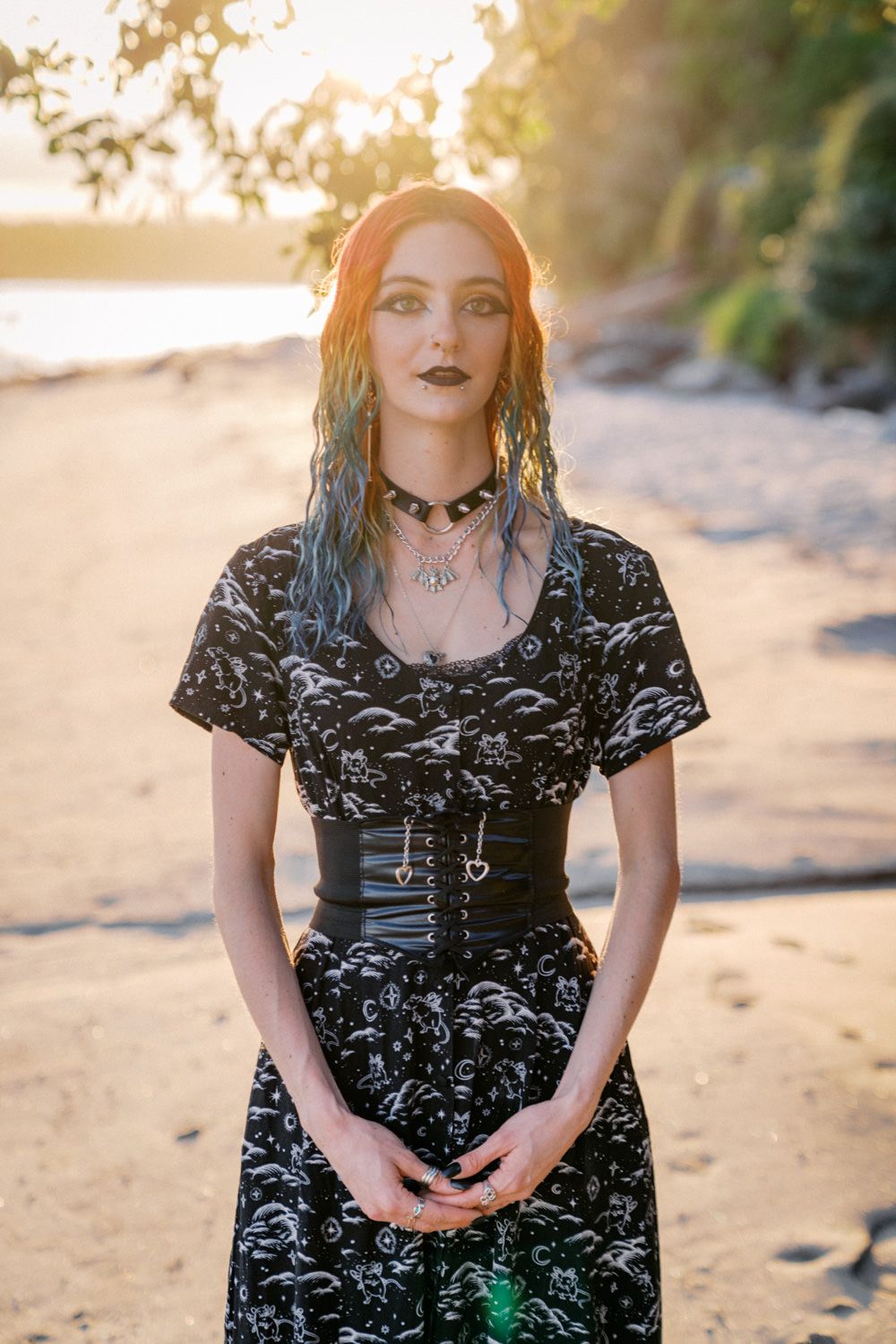Gothic woman with blue hair and black corset dress stands on a sandy beach at golden sunset.