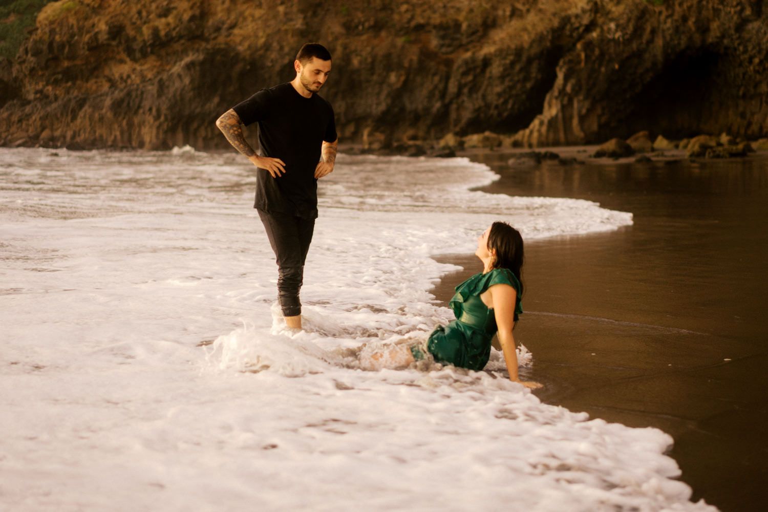 Couple enjoying ocean waves on a rocky beach, woman in green dress sitting in surf while tattooed man stands nearby.