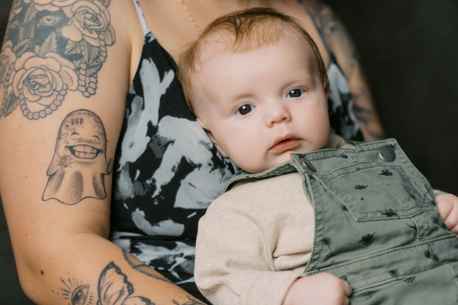 Tattooed parent holding an alert baby dressed in green overalls and a cream onesie.