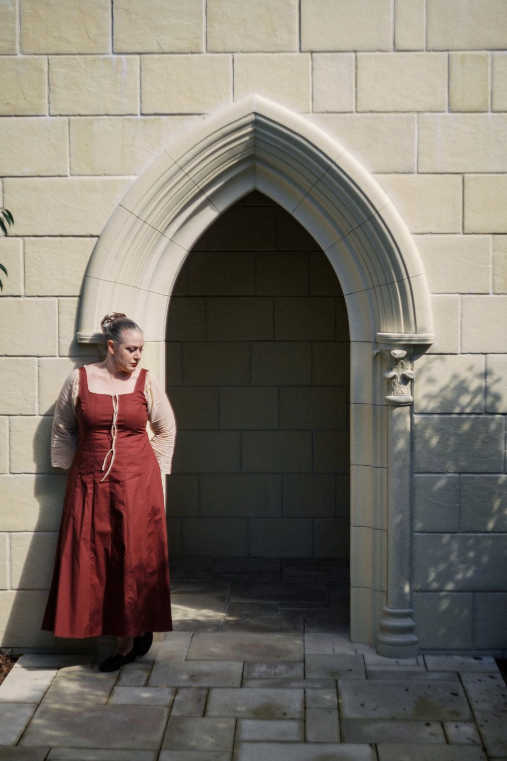 Woman in medieval red dress standing beside Gothic stone arch doorway of castle wall.