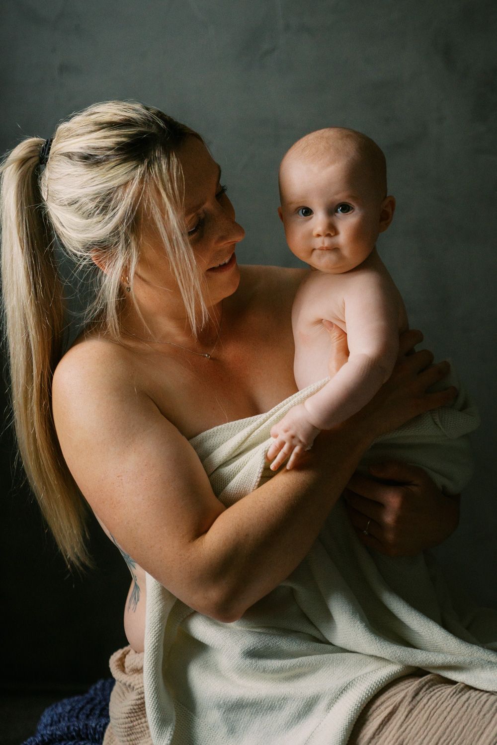 A blonde mother wrapped in a white towel tenderly holds her baby infant against a dark studio background.