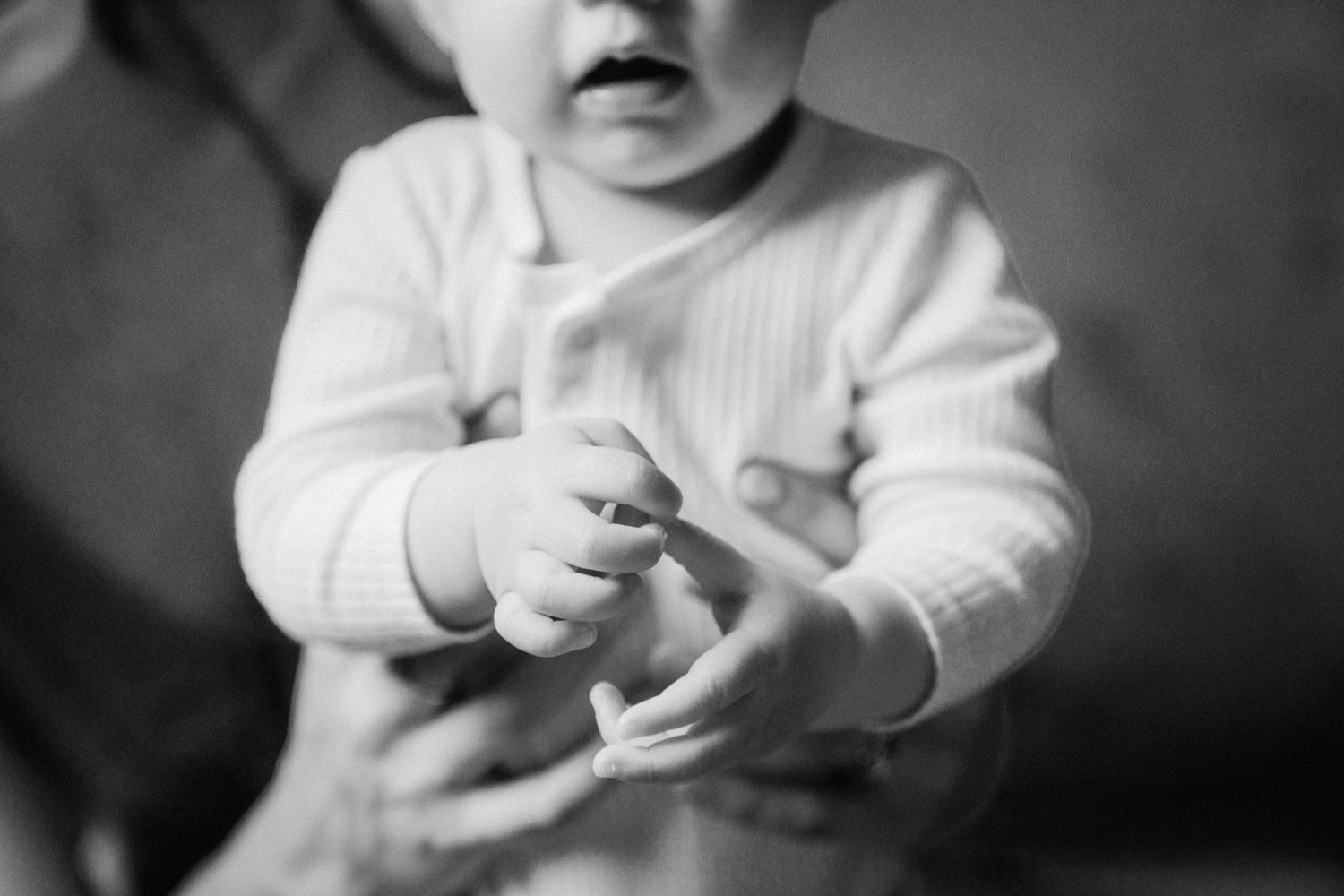 Black and white photo of a curious baby in a white onesie reaching out with tiny hands.