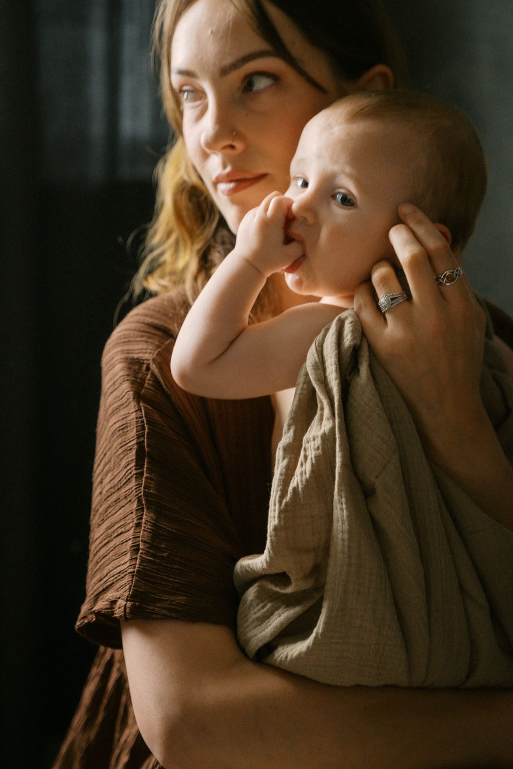 A mother tenderly holds her baby wrapped in a green muslin cloth, both bathed in warm natural light.