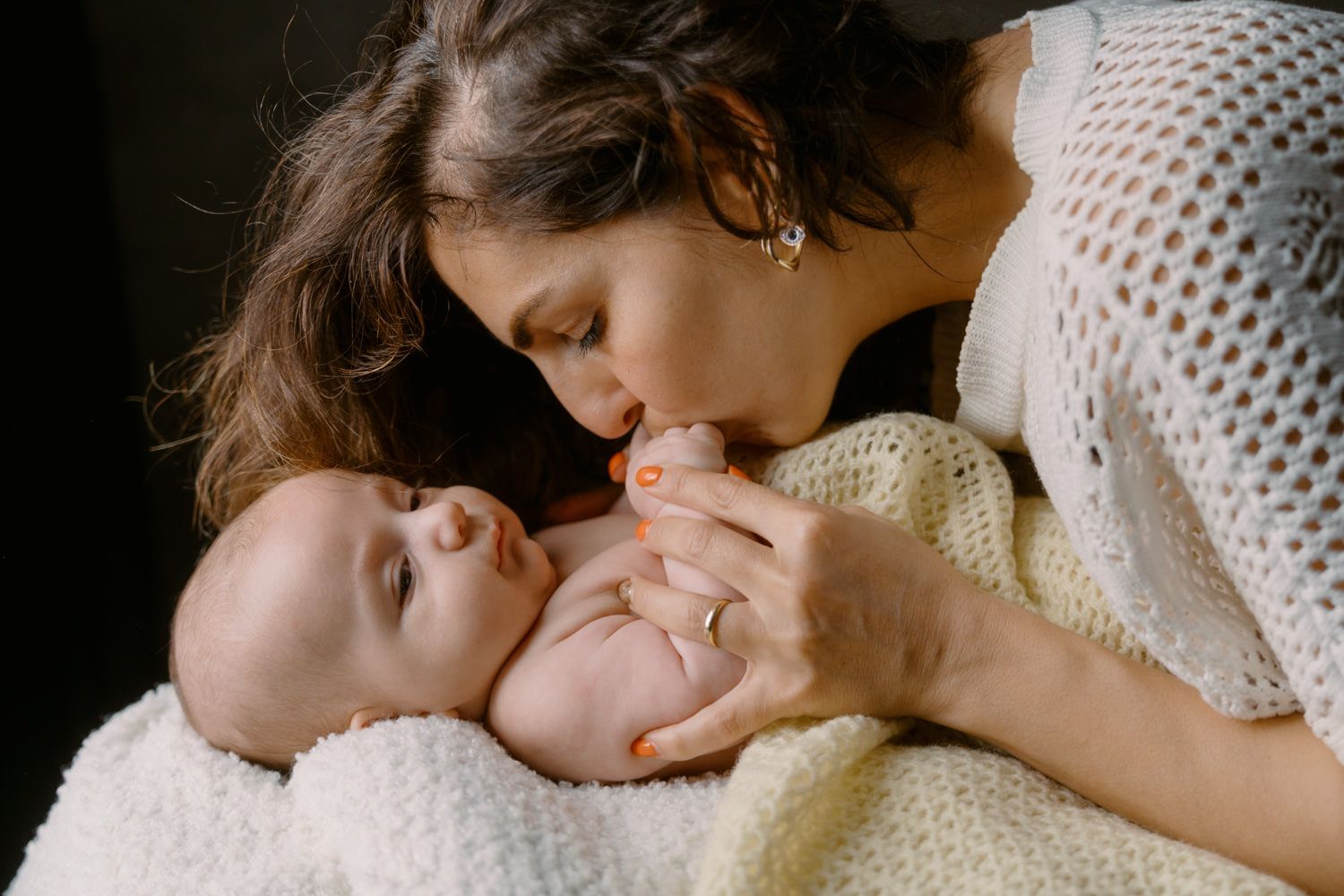 A loving mother gently kisses her newborn baby wrapped in a soft cream knit blanket.