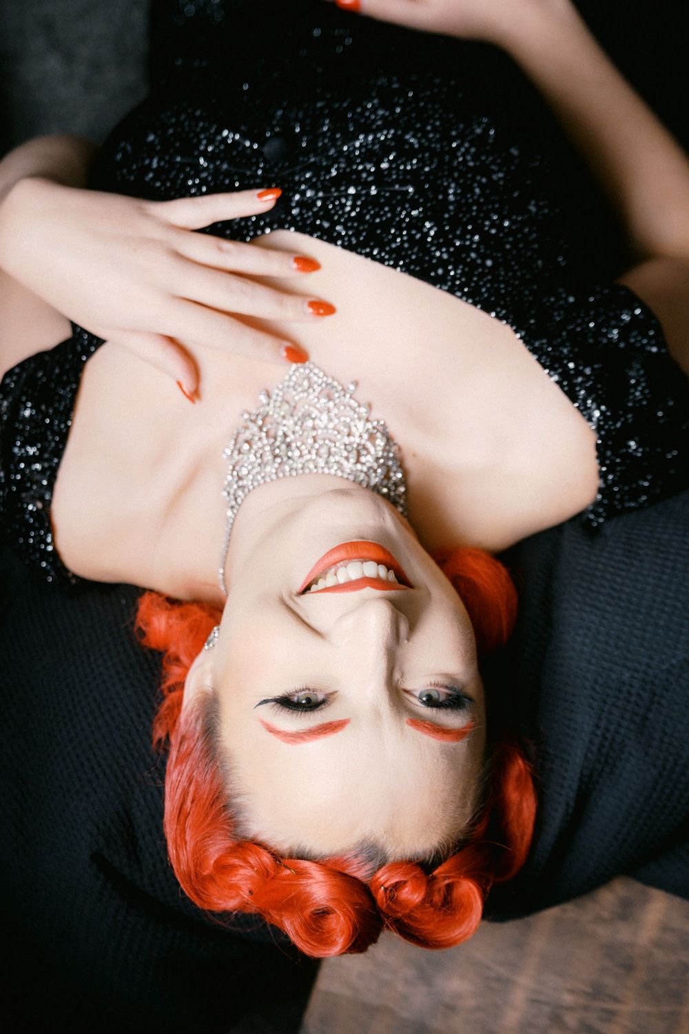 Woman with red hair and silver crown wearing black sequin dress, smiling while lying down.