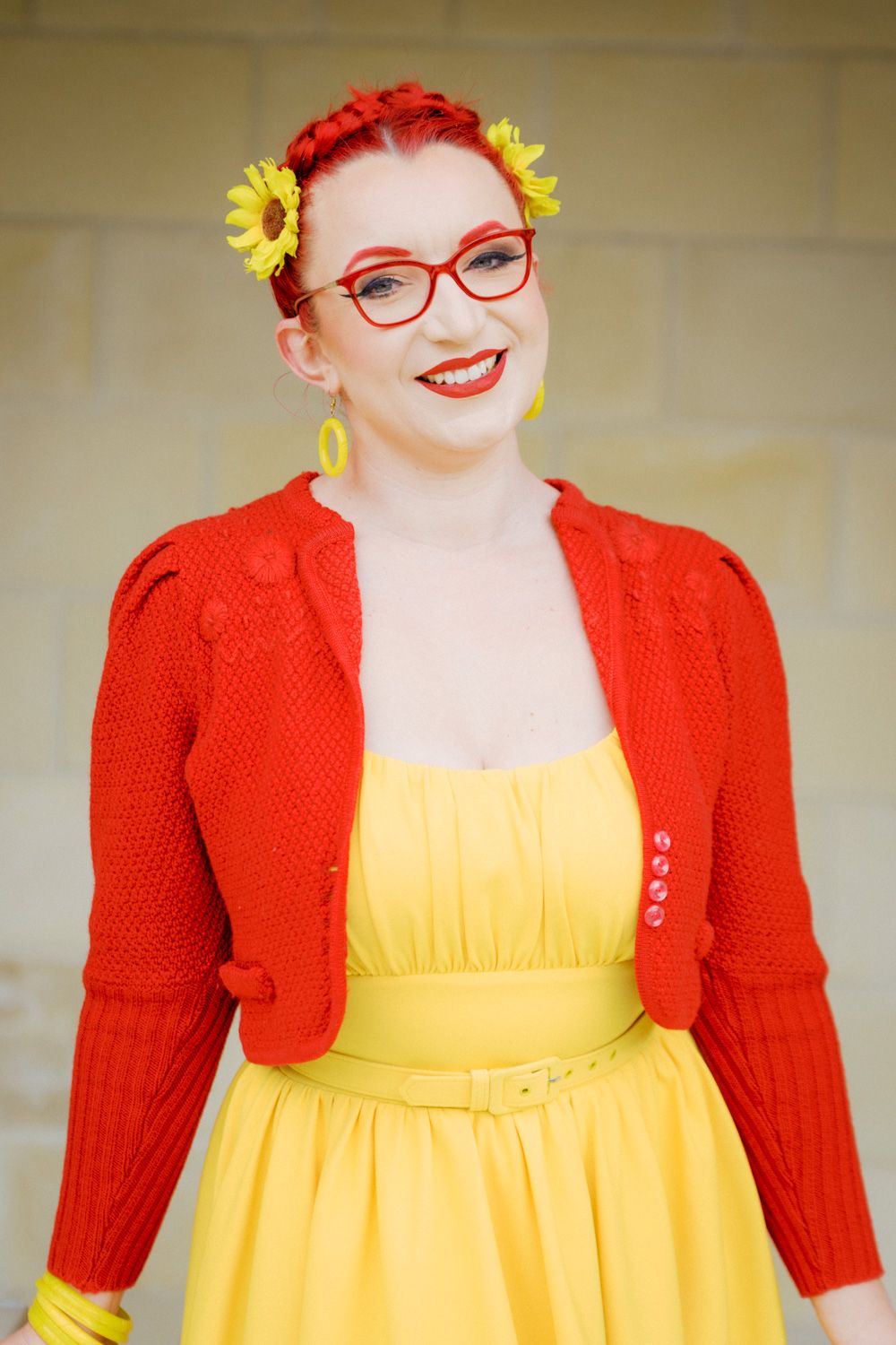 Smiling woman with red hair and glasses wearing a yellow dress and red cardigan with sunflower accessories.