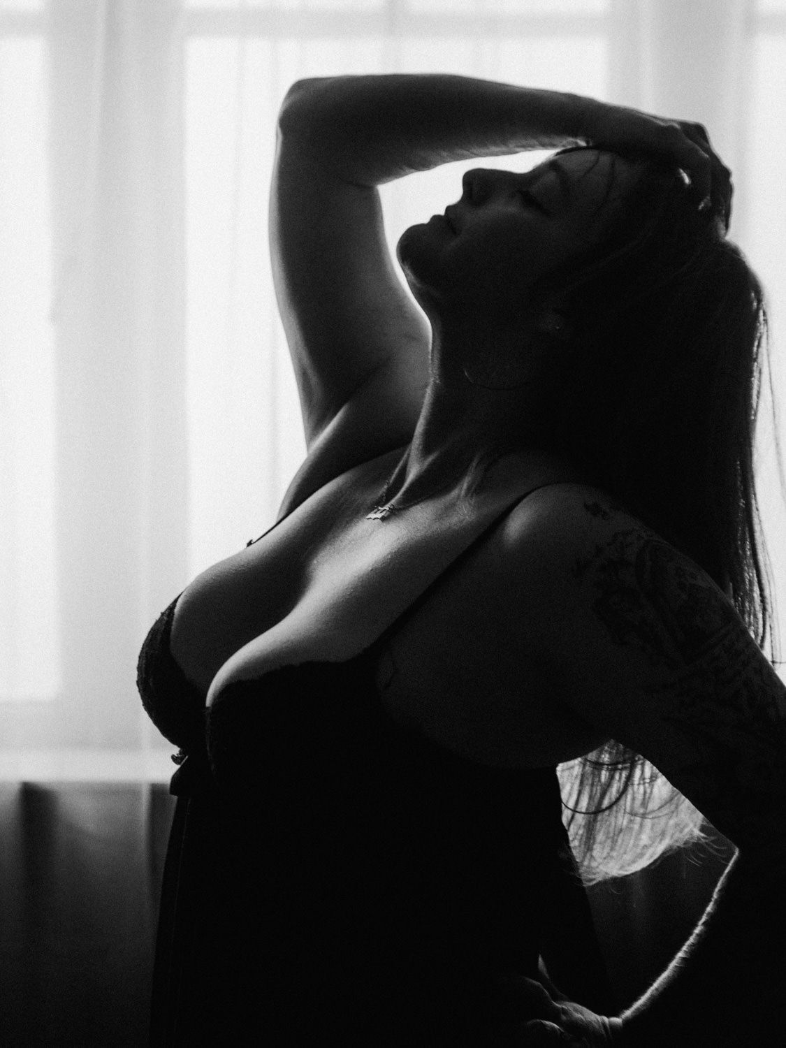 Black and white boudoir silhouette of a woman in lingerie posing gracefully against a bright window.