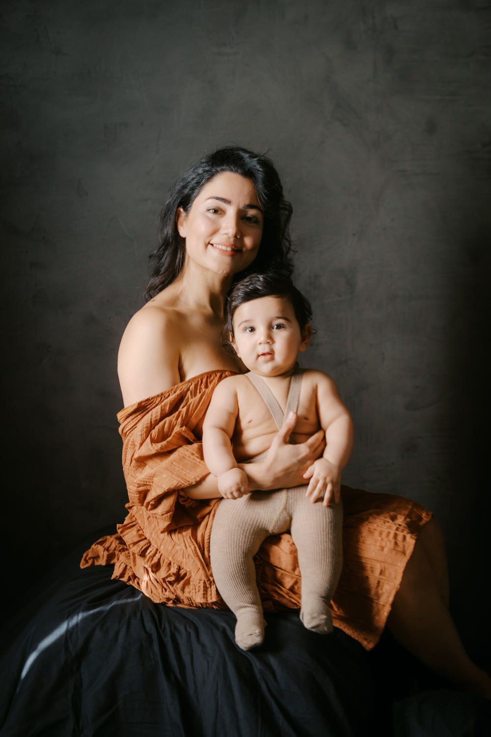 A smiling dark-haired woman in a rust orange wrap holds an adorable baby in a neutral onesie for a studio portrait.