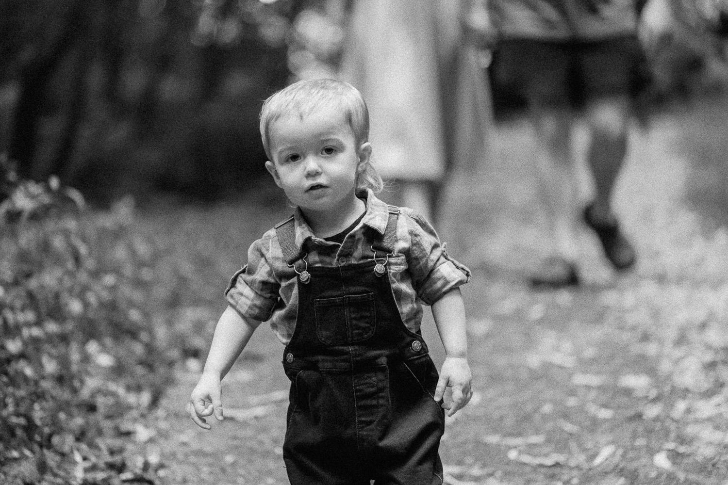 Black and white photo of a toddler boy in overalls and plaid shirt walking outdoors on a garden path.