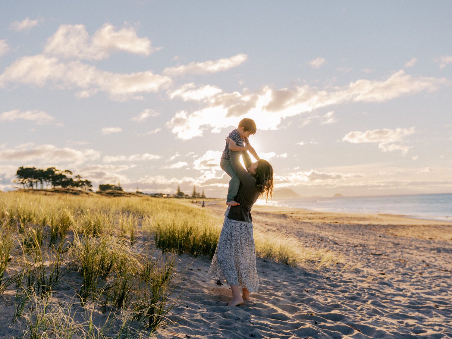 A parent joyfully lifts a child on a sunny beach with golden grass and ocean views at sunset.